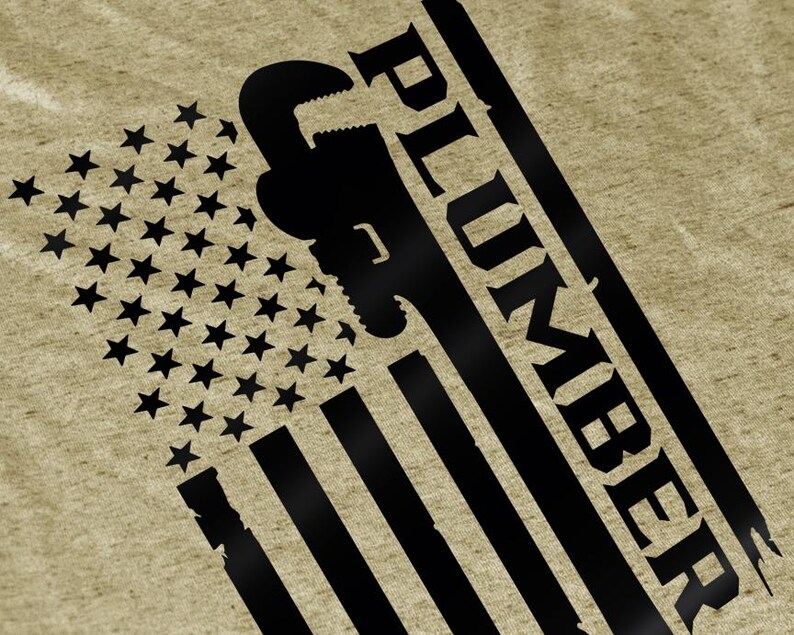 May include: A tan-coloured graphic tee featuring a distressed American flag design with a black wrench silhouette and the word "PLUMBER" in bold, angled text. The flag's stars and stripes are prominently displayed.
