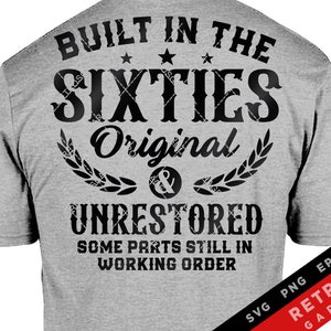 Peut inclure: Un t-shirt gris avec un texte noir qui dit "Built in the Sixties Original & Unrestored Some Parts Still in Working Order". Le texte est dans un style de police distressé. Le t-shirt a également un petit drapeau américain sur la manche gauche.