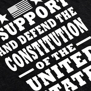 May include: Black t-shirt with white text that reads "I support and defend the Constitution of the United States". A small American flag is in the top left corner.