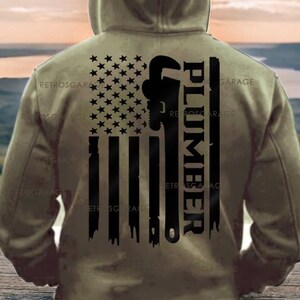 May include: A khaki hoodie featuring a distressed American flag design. The flag incorporates a wrench and the word "PLUMBER" in black. The hoodie has a hood and is viewed from the back.