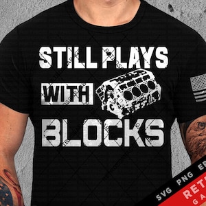 May include: Black t-shirt with white text that reads "Still Plays With Blocks". The text is in a bold, blocky font. A stylized image of an engine block is behind the word "With".