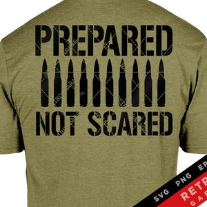 Puede incluir: Camiseta verde oliva con el texto "PREPARED NOT SCARED" en letras negras. Debajo del texto hay ilustraciones de balas negras. Una bandera estadounidense está en la manga izquierda.