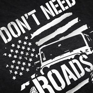 May include: Black and white graphic design on a dark grey t-shirt. The design features a jeep with the text "I don't need roads" and an American flag.