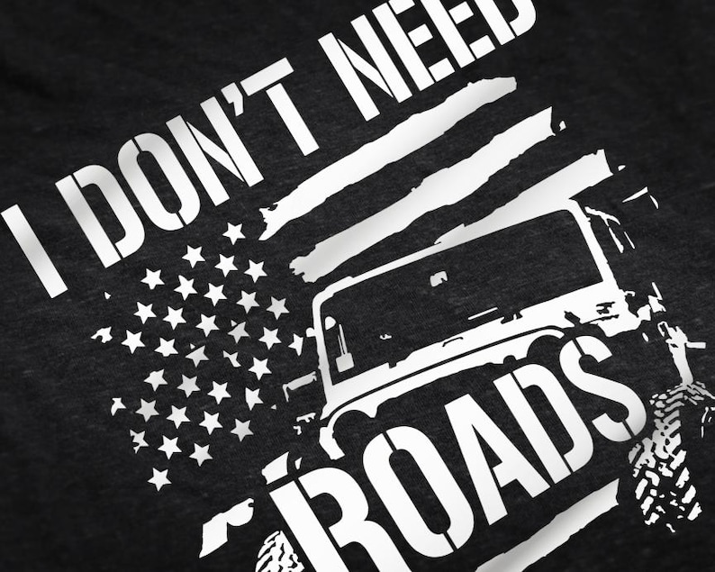 May include: Black and white graphic design on a dark grey t-shirt. The design features a jeep with the text "I don't need roads" and an American flag.