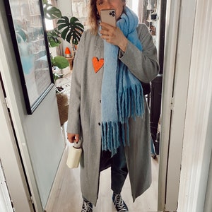 May include: A person wearing a long grey coat, a light blue scarf, and a grey sweater with an orange heart. They are holding a phone and a white water bottle. They are wearing dark blue trainers with white soles.