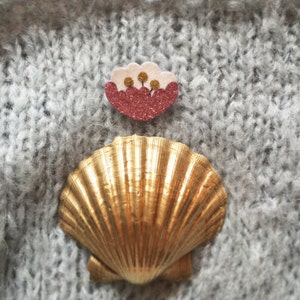May include: A gold seashell with a pink and white flower embellishment. The seashell is on a gray knitted fabric.