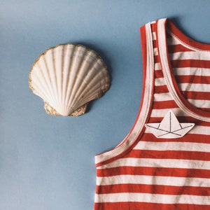 May include: A seashell with gold glitter trim and a red and white striped tank top with a paper boat applique on a light blue background. The tank top has white trim.