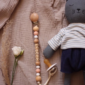 Op de afbeelding: Een houten baby-bijtje met een clip, kralen en houten blokken die de naam "SOPHIA" spellen. De bijtje ligt op een lichtbruin, getextureerd oppervlak met een grijs knuffeldier in de vorm van een kat.