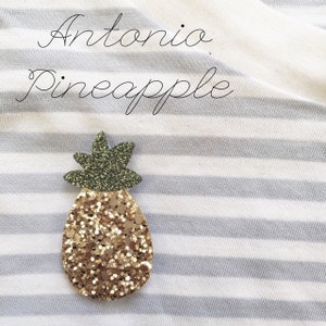 May include: A gold glitter pineapple with green glitter leaves. The pineapple is on a white and grey striped fabric. The text "Antonio Pineapple" is written above the pineapple.
