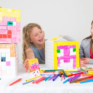 May include: Two children are colouring cardboard boxes with faces drawn on them, inspired by the popular video game Minecraft. The boxes are stacked and feature pixelated designs in shades of yellow, pink, orange, and white. The children are using colourful markers to add details to the faces.
