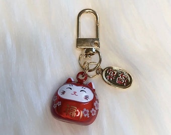 May include: A red and gold lucky cat keychain with a small charm that says "Good Fortune" in Japanese. The cat is holding a coin and has a happy expression.