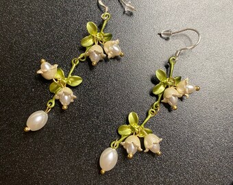 May include: A pair of floral earrings featuring a delicate design. The earrings have a gold-tone metal stem with green leaves and small, off-white bell-shaped flowers. Each earring has a dangling, pear-shaped pearl.