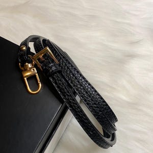 May include: Black leather strap with a gold metal clasp and a loop. The strap is folded in half and is lying on a black surface.