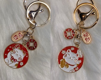 May include: Two gold keychains with red and white lucky cat charms. Each charm features a white cat with a gold coin in its paw and the Chinese character for "fortune" written in red. The keychains also include a gold coin charm with Chinese characters and a red tag with the Chinese character for "good fortune" written in gold.