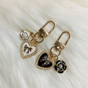 May include: Two gold keychains with heart-shaped charms. One charm is white with a black bow and the other is black with a white bow. Both charms have a pearl border and are accented with a small flower charm.