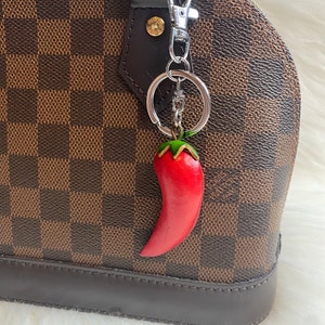 May include: A red chilli pepper keychain attached to a silver keyring. The keychain is hanging from a brown and tan checkered handbag.
