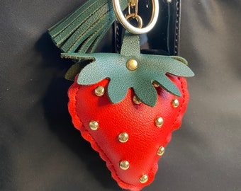 May include: A red leather strawberry keychain with gold studs and green leather leaves. The keychain is attached to a gold key ring.