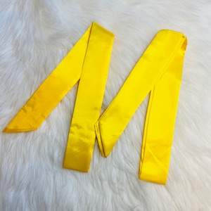 May include: Two yellow satin ribbon sashes, folded and arranged in an M shape on a white fluffy surface.