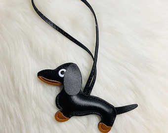 May include: Black leather dachshund keychain with brown accents and white eyes. The keychain has a black leather loop for attaching to a bag or keys.