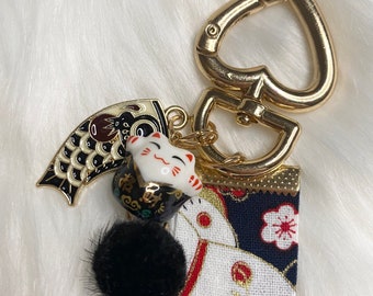 May include: A gold heart-shaped keychain with a black and white fabric charm featuring a Japanese lucky cat design, a black pom-pom, and a gold koi fish charm.