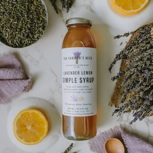 Puede incluir: Una botella de vidrio de jarabe simple de lavanda y limón, con tapa plateada, sobre una superficie de mármol. La etiqueta incluye el texto "handmade from our farm to you". La imagen incluye lavanda, rodajas de limón y una cuchara de madera.