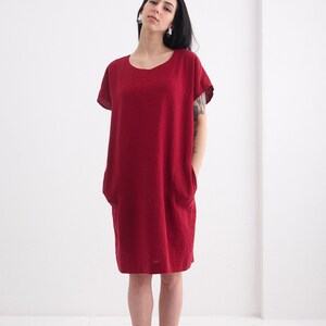 Puede incluir: Un vestido de lino burdeos con mangas cortas y cuello redondo. El vestido tiene dos bolsillos en la parte delantera y un corte holgado.