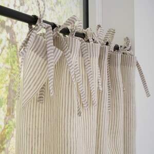Puede incluir: Cortinas a rayas color crema con cierres de lazo. Las rayas verticales son de color marrón claro. Las cortinas cuelgan de una barra negra frente a una ventana con vistas a árboles.