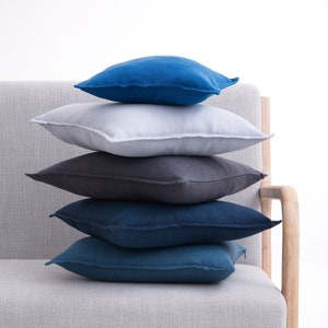 Stacked linen pillow covers in shades of blue, gray, and white on a light gray sofa with wooden armrests, against a white background.