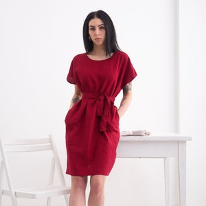 Puede incluir: Una mujer con un vestido de lino rojo con cintur&oacute;n de lazo y mangas cortas. Est&aacute; de pie frente a una mesa y una silla blancas.
