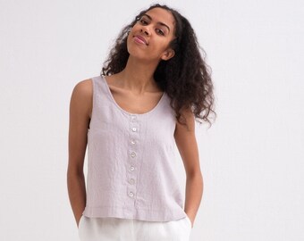 May include: A woman wearing a light purple sleeveless linen top with a button-up front. She is smiling and looking at the camera.