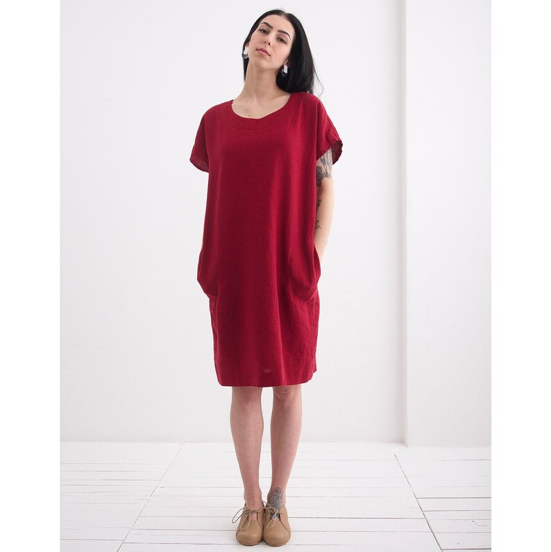 Puede incluir: Un vestido de lino burdeos con mangas cortas y cuello redondo. El vestido tiene dos bolsillos en la parte delantera y un corte holgado.