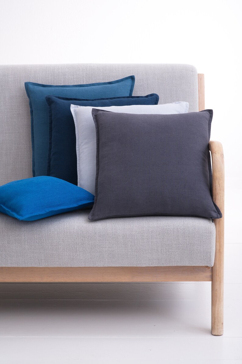 Modern light gray sofa adorned with various shades of blue and gray cushions, with a wooden armrest and base, set against a white background.