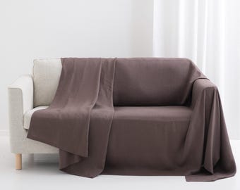 May include: A solid dark brown rectangular throw blanket draped over a light beige sofa. The fabric has a matte finish and hangs over the back, seat, and one arm of the sofa, creating soft folds and a smooth, consistent texture across the surface.