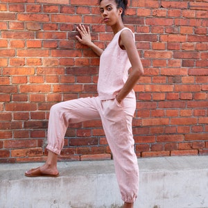 Op de afbeelding: Een vrouw draagt een roze linnen broekpak met een mouwloos topje en getailleerde broek. Ze staat met &eacute;&eacute;n voet op een betonnen rand en de andere voet op de grond. Ze kijkt naar de camera.