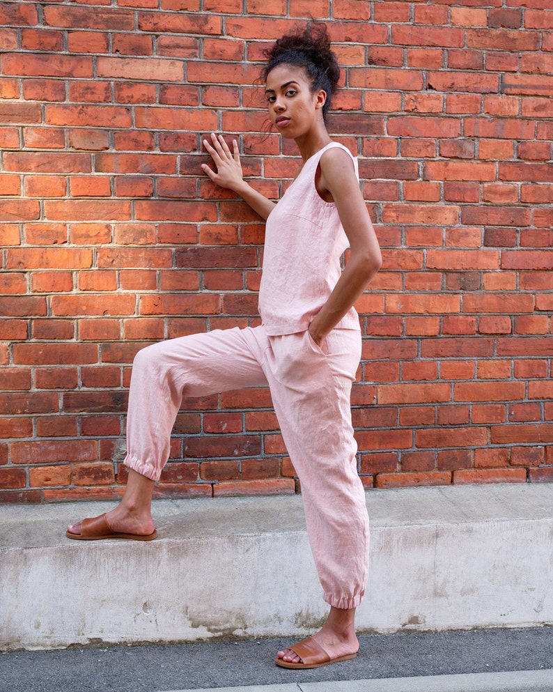 Op de afbeelding: Een vrouw draagt een roze linnen broekpak met een mouwloos topje en getailleerde broek. Ze staat met &eacute;&eacute;n voet op een betonnen rand en de andere voet op de grond. Ze kijkt naar de camera.