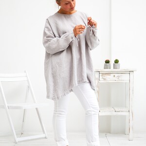 May include: A woman wearing a light grey linen tunic top with long sleeves and white jeans. The sleeves have ties at the wrists. She is standing on a white wooden floor and is looking down at her hands.