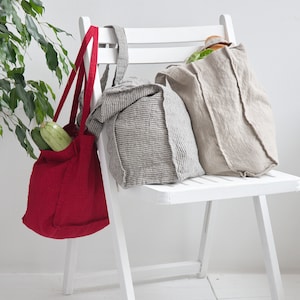 May include: Three reusable shopping bags hanging on a white chair. The bags are made of linen and have a natural, rustic look. One bag is red, one is gray and white striped, and one is beige. The red bag has two zucchinis inside.