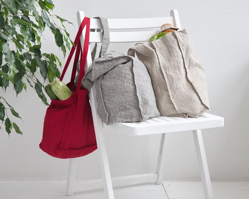 May include: Three reusable shopping bags hanging on a white chair. The bags are made of linen and have a natural, rustic look. One bag is red, one is gray and white striped, and one is beige. The red bag has two zucchinis inside.