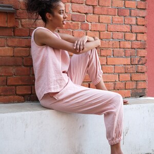Op de afbeelding: Een vrouw in een roze top zonder mouwen en een bijpassende broek zit op een witte rand. Ze draagt ​​bruine sandalen en heeft haar haar in een knotje.