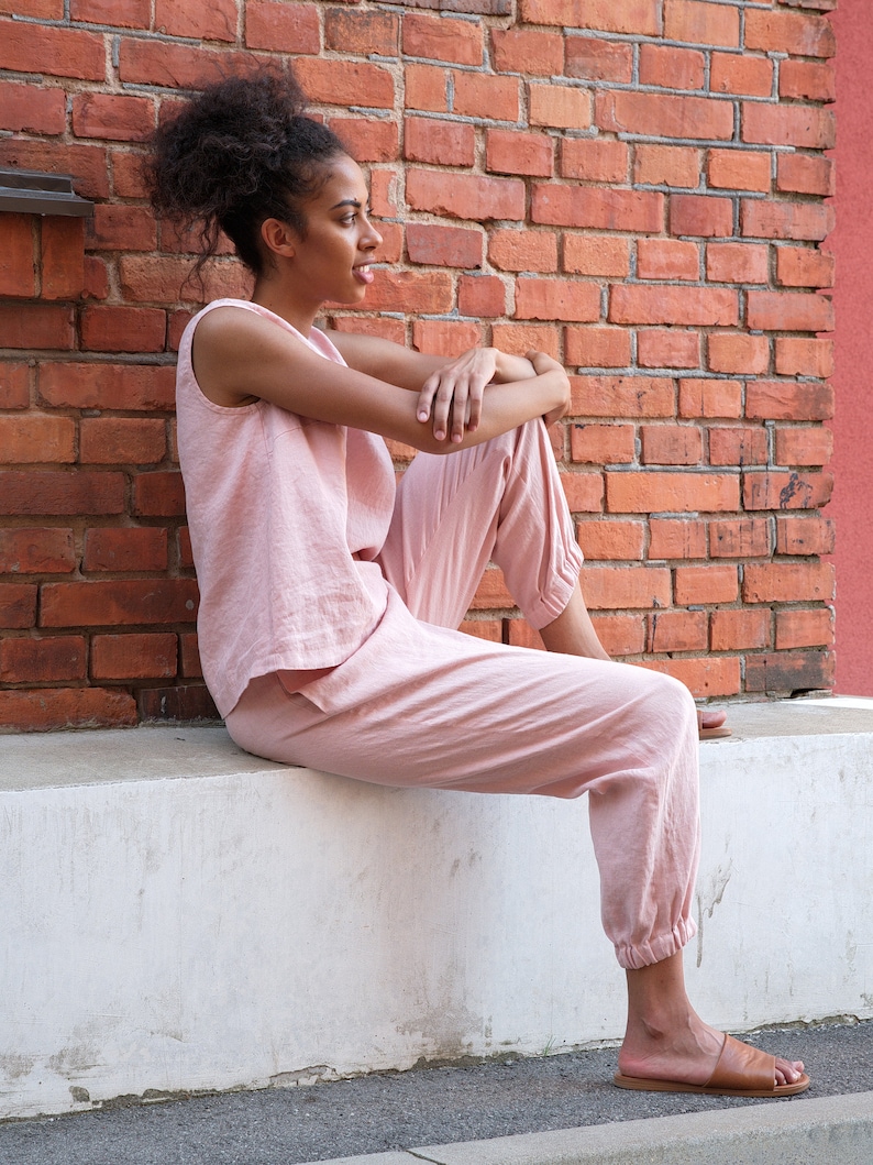 Op de afbeelding: Een vrouw in een roze top zonder mouwen en een bijpassende broek zit op een witte rand. Ze draagt ​​bruine sandalen en heeft haar haar in een knotje.