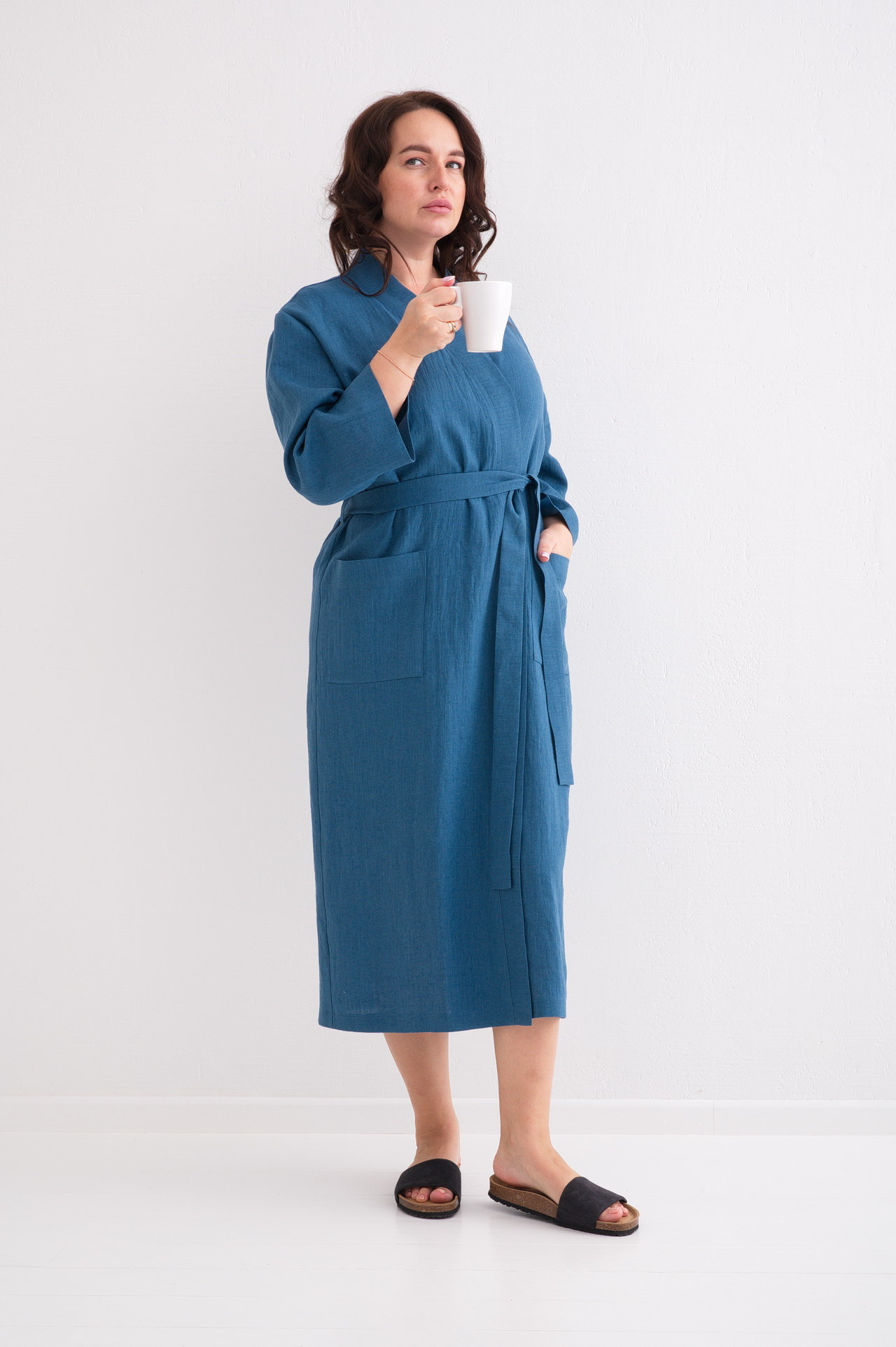 May include: A woman wearing a blue linen robe with a white belt and pockets. She is holding a white mug in her right hand and standing on a white background.