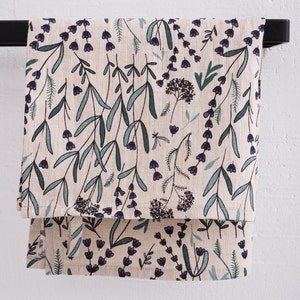 Peut inclure: Une serviette de cuisine en lin beige avec un motif floral de feuilles vertes et de fleurs violettes. La serviette est suspendue &agrave; un porte-serviettes noir.