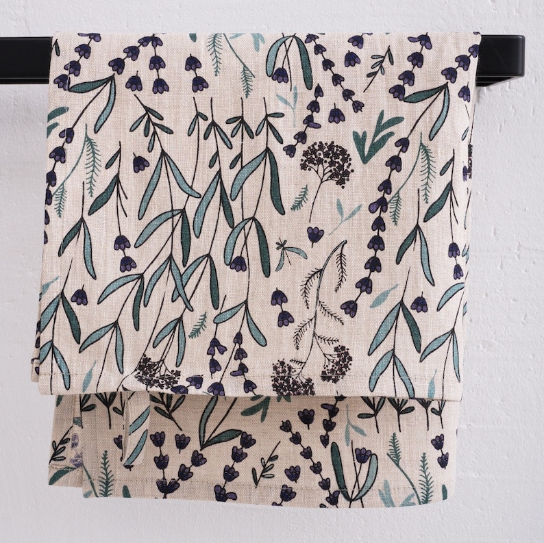 Peut inclure: Une serviette de cuisine en lin beige avec un motif floral de feuilles vertes et de fleurs violettes. La serviette est suspendue &agrave; un porte-serviettes noir.