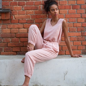 Op de afbeelding: Een vrouw draagt een lichtroze, mouwloze jumpsuit met een v-hals en een trekkoord in de taille. Ze zit op een witte rand met gekruiste benen en draagt ​​bruine sandalen.