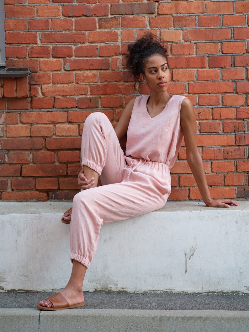 Op de afbeelding: Een vrouw draagt een lichtroze, mouwloze jumpsuit met een v-hals en een trekkoord in de taille. Ze zit op een witte rand met gekruiste benen en draagt ​​bruine sandalen.