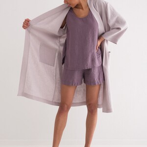 Puede incluir: Conjunto de bata, camiseta sin mangas y pantalones cortos de lino lila. La bata tiene bolsillos grandes y un corte holgado. La camiseta sin mangas y los pantalones cortos son de un tono p&uacute;rpura m&aacute;s oscuro. La persona lleva chanclas negras.