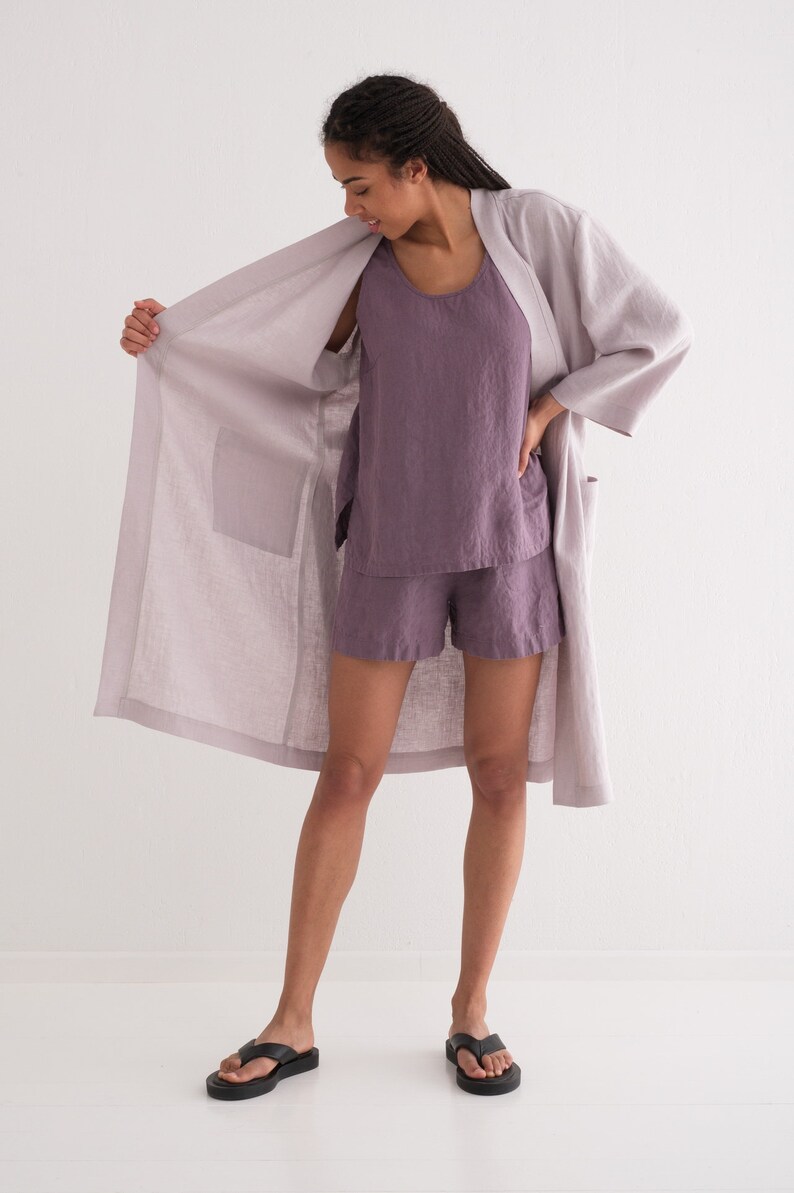 Puede incluir: Conjunto de bata, camiseta sin mangas y pantalones cortos de lino lila. La bata tiene bolsillos grandes y un corte holgado. La camiseta sin mangas y los pantalones cortos son de un tono p&uacute;rpura m&aacute;s oscuro. La persona lleva chanclas negras.