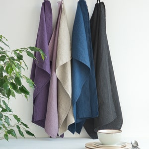 May include: Four linen kitchen towels hanging on hooks. The towels are in shades of purple, beige, blue, and black.