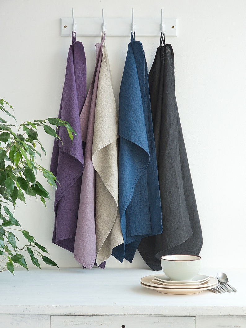 May include: Four linen kitchen towels hanging on hooks. The towels are in shades of purple, beige, blue, and black.