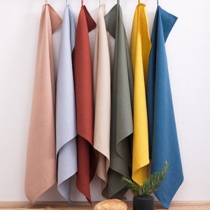Colorful linen towels in shades of beige, blue, rust, olive, and yellow, hanging on a black metal rack against a white wall with a wooden surface below, where a loaf of bread and a potted plant are placed.